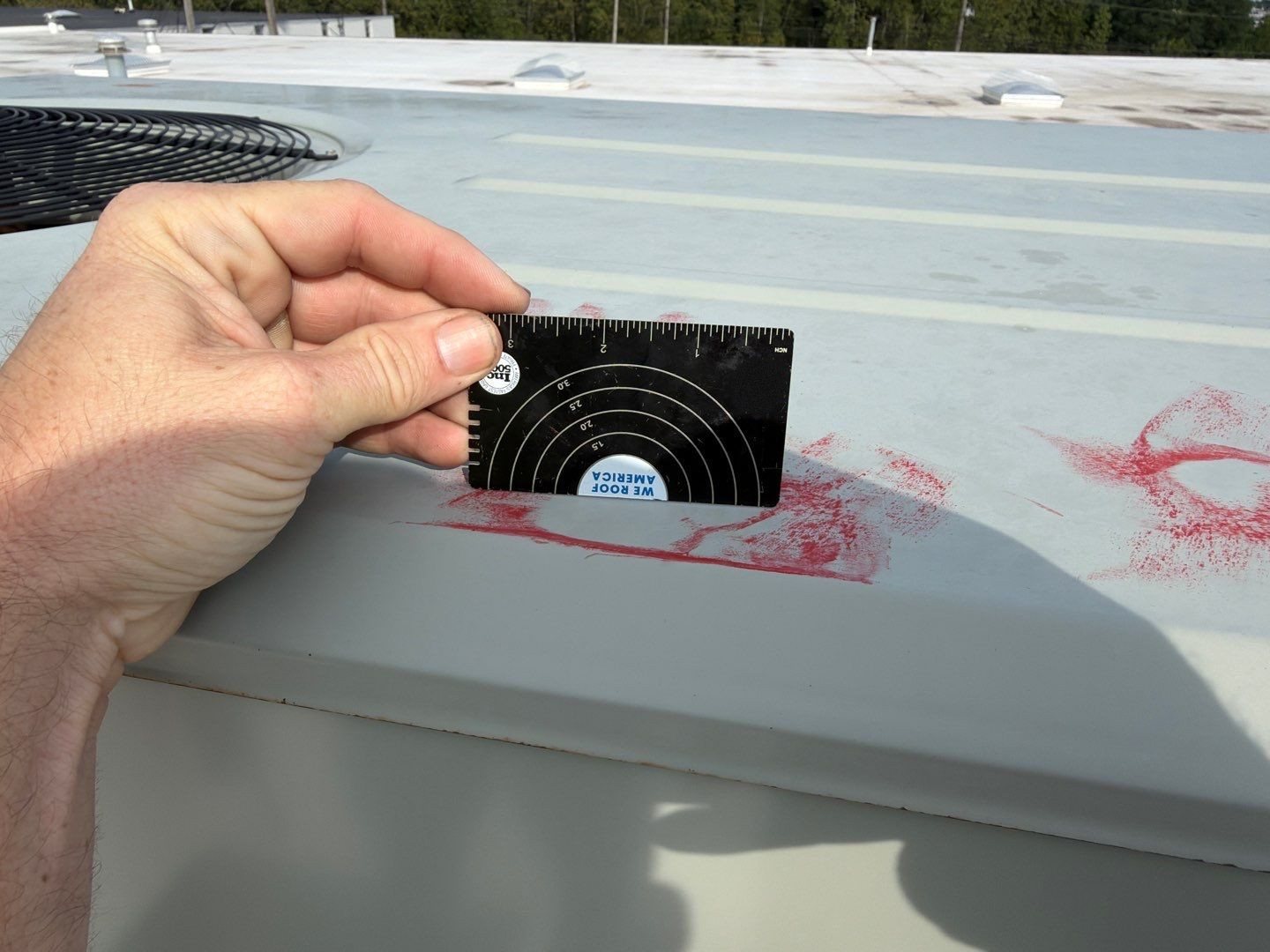 A hail gauge held next to a marked impact on a TPO roof